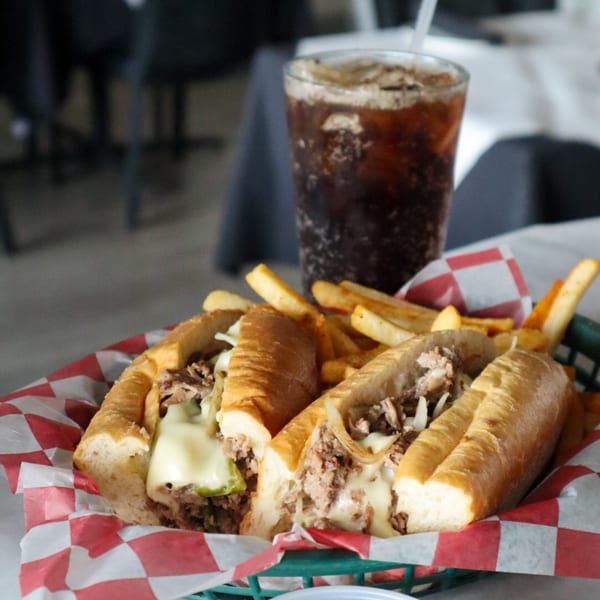 Two Italian cheesesteak sandwiches with fries in a basket, and a soda in the background.