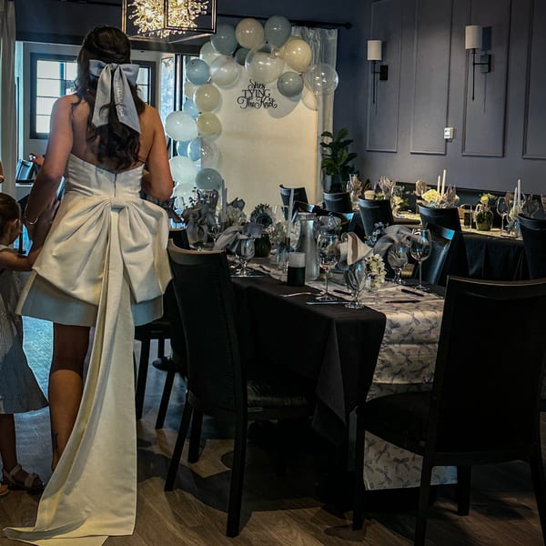 Woman in a white dress with a large bow enters a private dining room set for a celebration.