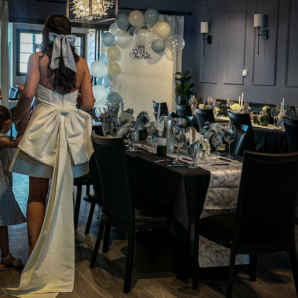 Woman in a white dress with a large bow enters a private dining room set for a celebration.