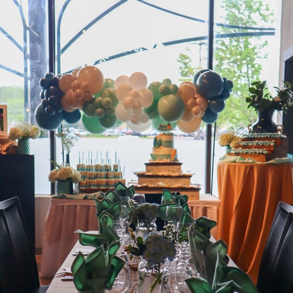 A decorated dining table with green napkins, glasses, and floral centerpieces, with cakes and balloons in the background.